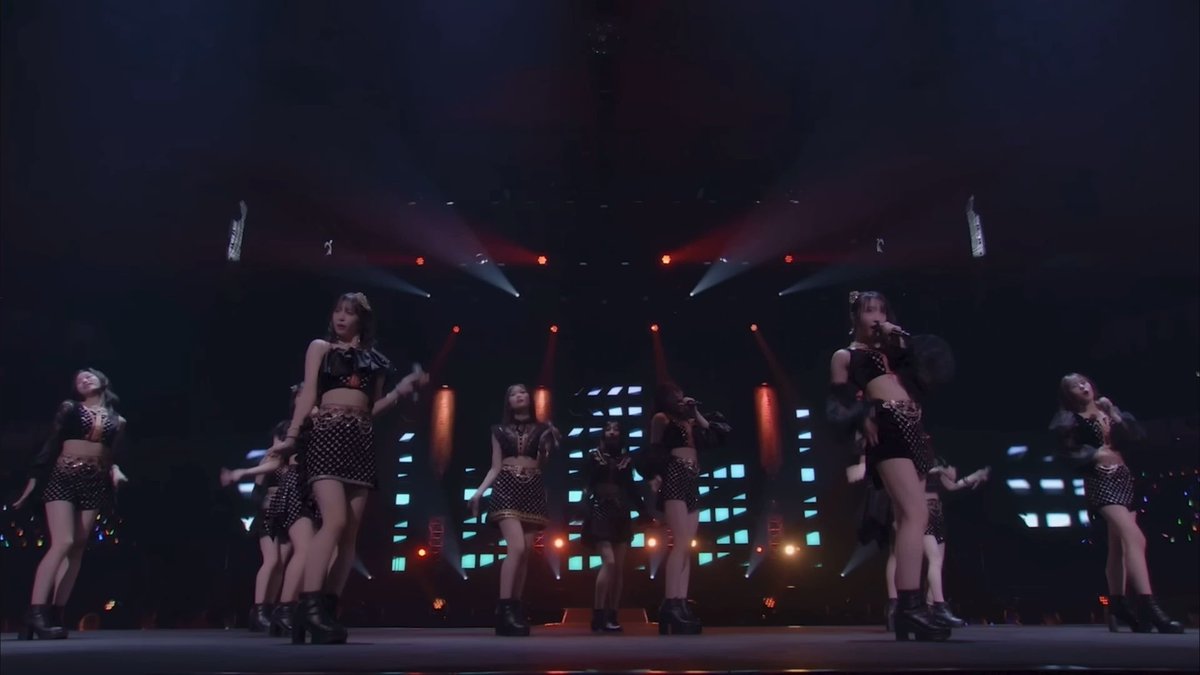 A group of female performers in matching black studded crop tops and skirts dance on a dimly lit stage illuminated by spotlights and LED screens.