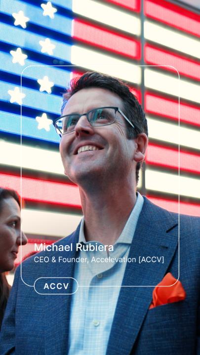 Michael Rubiera, CEO & Founder of Accelevation [ACCV], smiles during a Nasdaq appearance, framed against the iconic American flag backdrop of the exchange's digital display. The on-screen caption "ACCV" reflects the company's milestone of being featured on Nasdaq, tying into his remarks about the generational build-out of America's digital economy and manufacturing innovation.