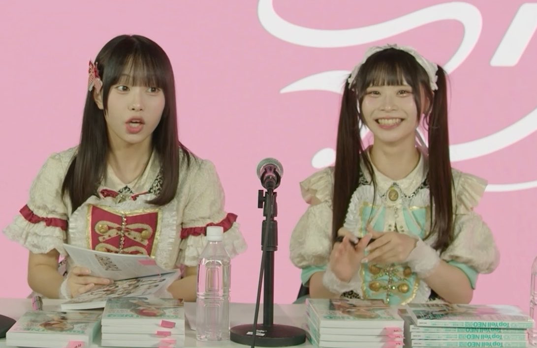 Two young women in frilly pastel outfits sit at a table with a microphone and stacks of booklets during what appears to be a press event, with pink-themed books visible.