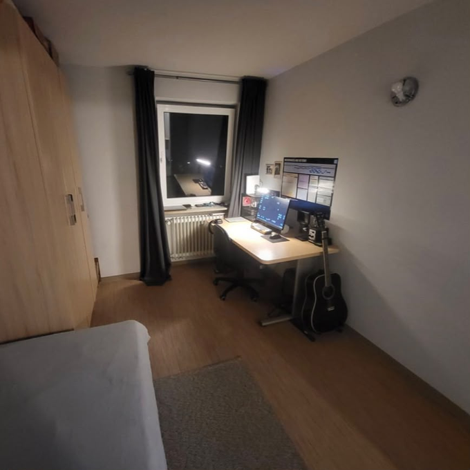 A dimly lit bedroom interior at night featuring a wooden desk with dual monitors displaying charts, a window with curtains, a guitar leaning against the wall, a wardrobe, and a bed in the foreground.