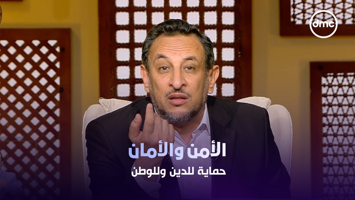 A man in a dark suit speaking in a studio setting with wooden lattice backgrounds, featuring Arabic text reading 'الأمن والأمان حماية للدين والوطن' and a 'dmc' logo.