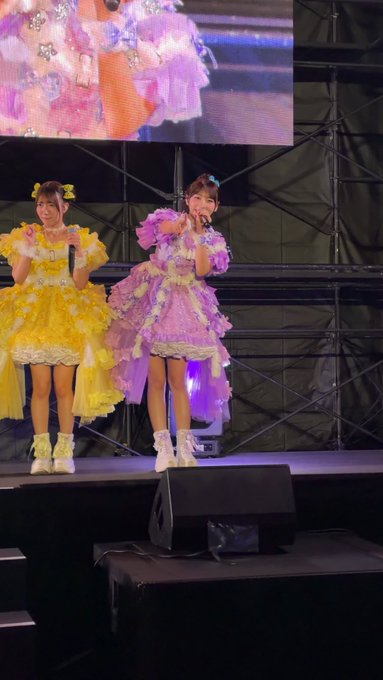Two performers in elaborate yellow and pink frilly dresses with matching bows perform on stage, with a large screen behind them.