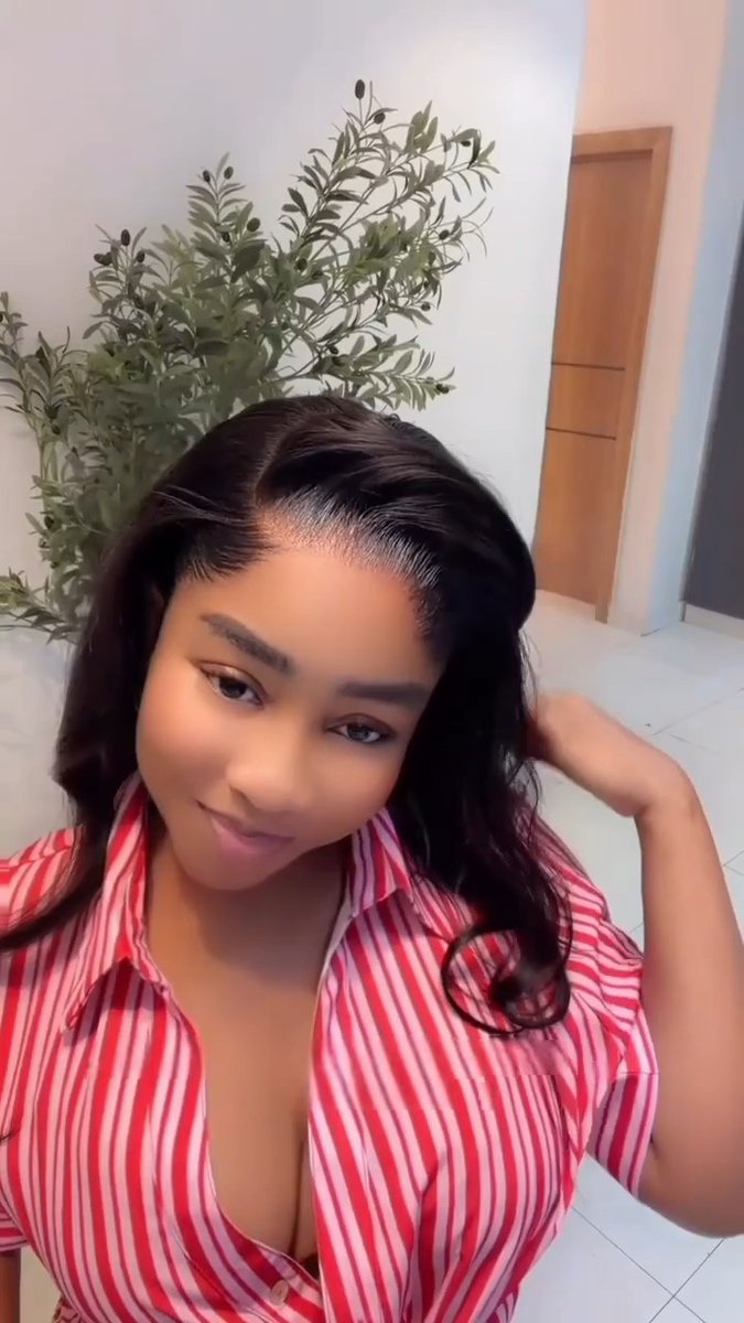 A woman with long dark wavy hair wearing a red and white vertically striped button-up shirt takes a selfie, with her hand raised behind her head, set against a backdrop of a leafy plant and wooden door. The image conveys a hopeful, contemplative mood that complements her prayer for things to work out.