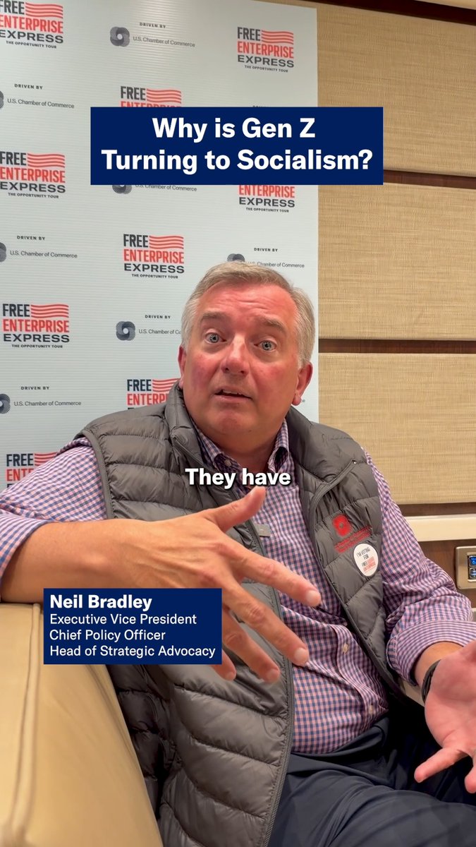 A man wearing a dark vest with a "Free Enterprise Express" badge speaks in front of a branded backdrop, with on-screen text identifying him as Neil Bradley, Executive Vice President, Chief Policy Officer, and Head of Strategic Advocacy. The caption "Why is Gen Z Turning to Socialism?" appears at the top, with "They have" displayed mid-video, aligning with his message that young people's frustrations stem from government interference in housing, healthcare, and higher education.