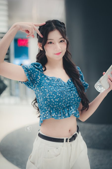 A young woman with long dark hair poses in a blue floral crop top and white pants, holding a water bottle and making a peace sign near her head.