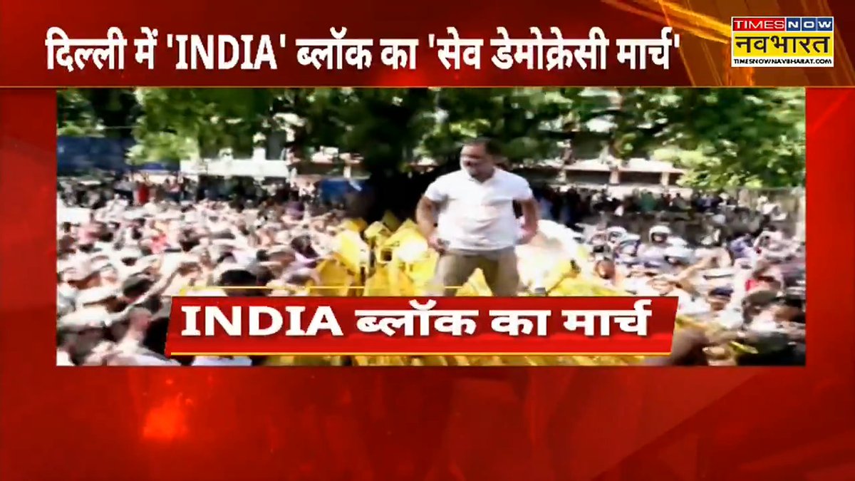 A Times Now Navbharat news broadcast screenshot with Hindi headline 'दिल्ली में INDIA ब्लॉक का सेव डेमोक्रेसी मार्च' (INDIA bloc's Save Democracy March in Delhi), showing a man in a white shirt surrounded by a large crowd of supporters.