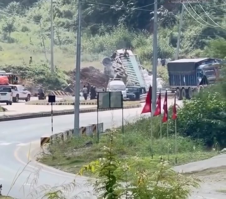 An outdoor scene showing an overturned truck on a roadside with smoke, surrounded by other vehicles, red flags, and people on a hilly road.