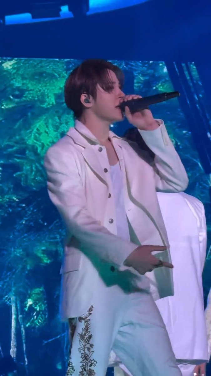 A male performer singing into a handheld microphone on stage, wearing a white jacket and white pants with floral embroidery, with a green forest-themed backdrop behind him.