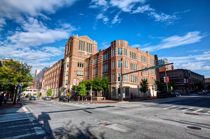 Large historic brick building on a sunny city street corner under a bright blue sky with scattered clouds.