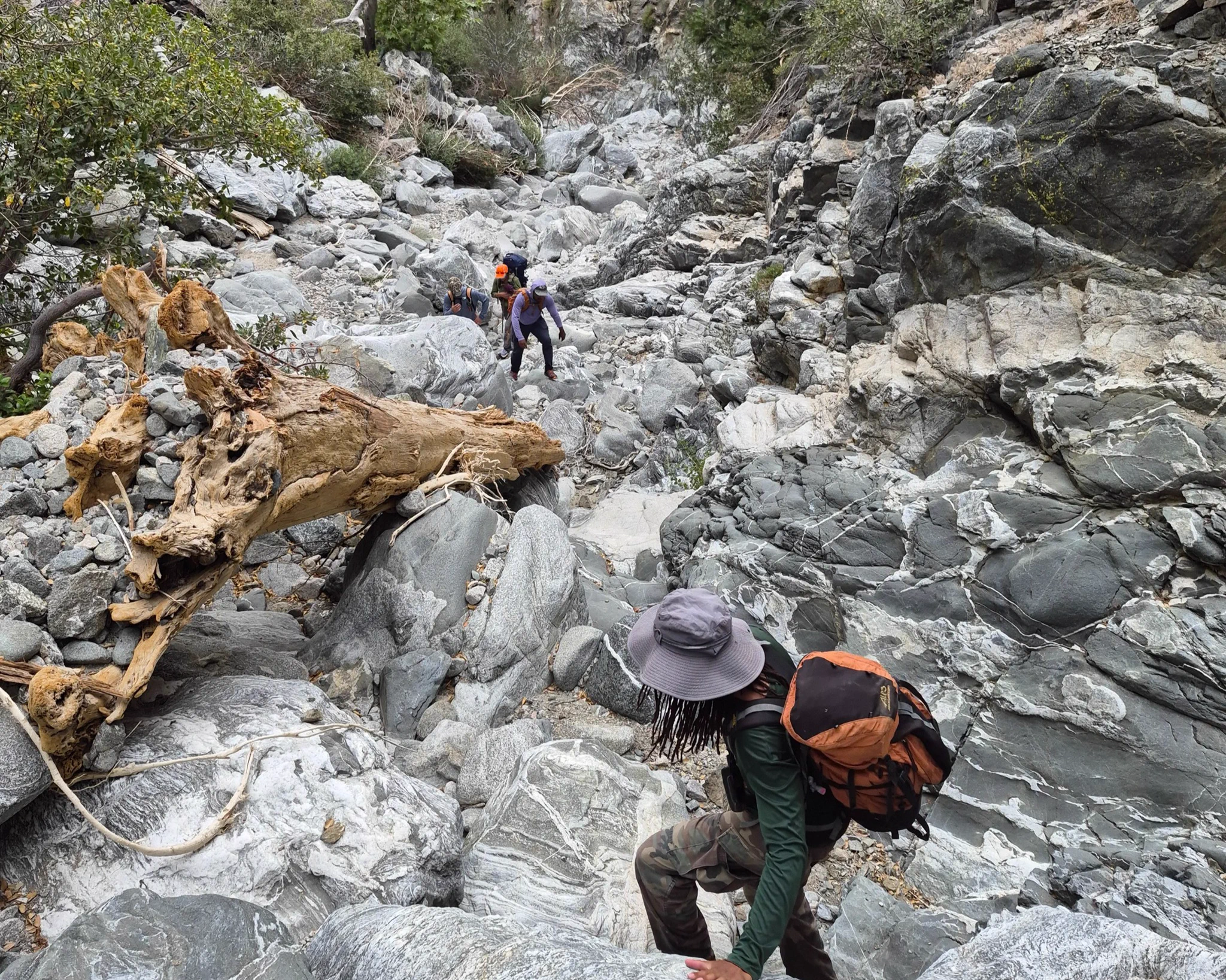 Three hikers navigate a rocky, steep mountain trail surrounded by large boulders and sparse vegetation.