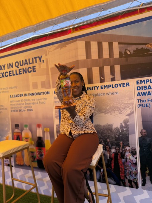 A smiling woman sits at an outdoor event booth holding up a bottle, with banners behind her reading 'A LEADER IN INNOVATION' and 'COMMUNITY EMPLOYER'.