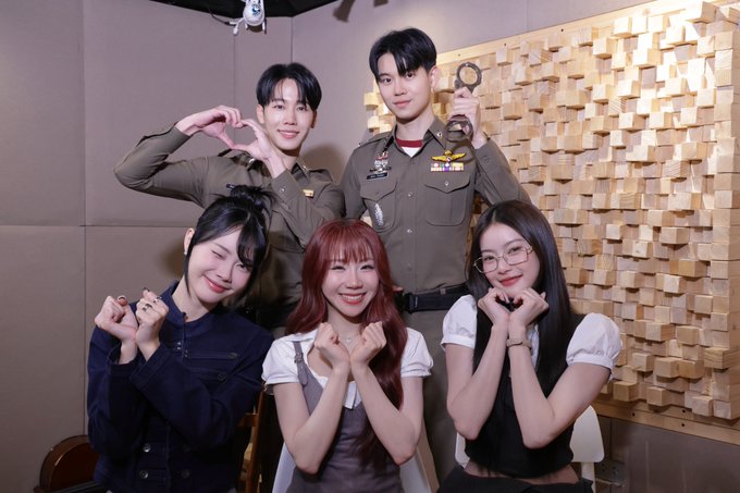 Five people pose together indoors, with two men in the back making heart and circle hand gestures in front of a wooden textured wall, while three women in front smile with clasped hands.