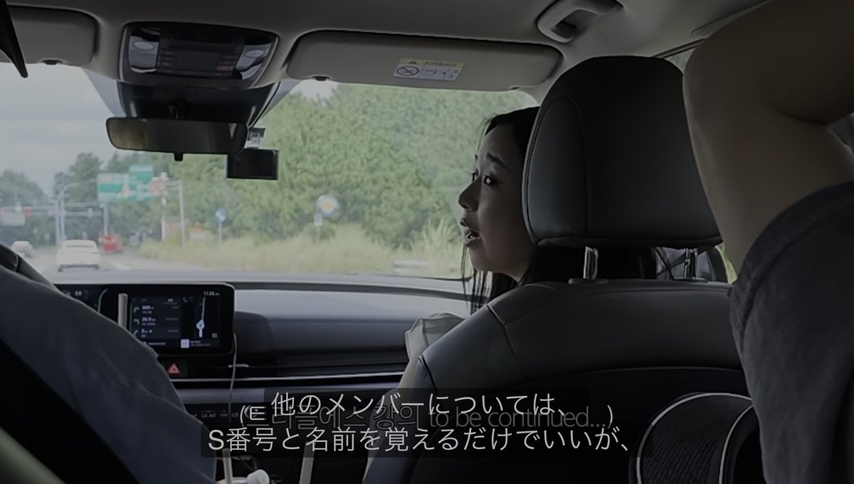 Inside a vehicle driving through a tree-lined area, a young woman with long dark hair sits in the back seat looking forward, with Japanese subtitles reading "他のメンバーについてはS番号と名前を覚えるだけでいいが" and a "(to be continued...)" caption overlaid on the scene.