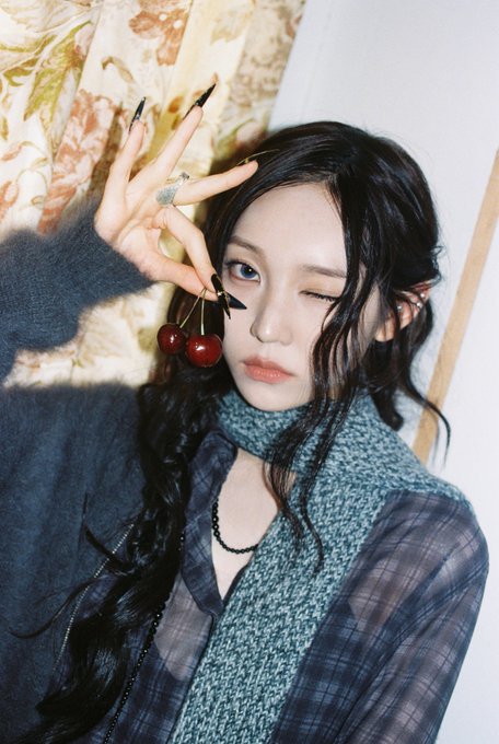 A young woman with long dark hair posing indoors, holding two cherries near her eye while wearing a plaid jacket and knit scarf.