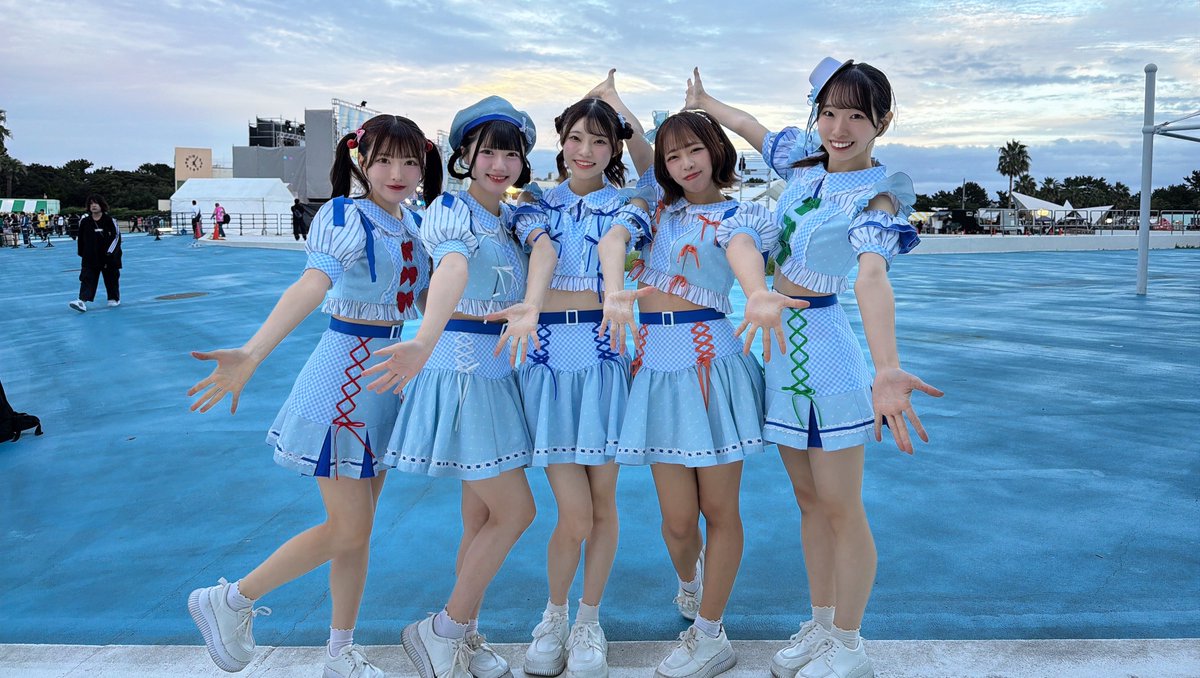 Five young women in matching light blue outfits with colorful accents pose together with their arms extended on a blue surface at dusk.