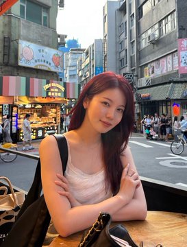 A young woman with long reddish hair sits at an outdoor cafe table with her arms crossed, with a busy city street and shops visible behind her.
