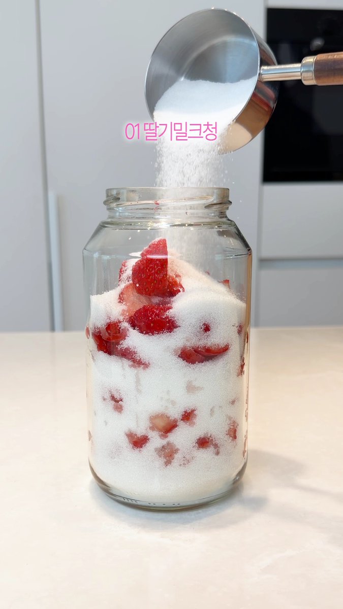 A clear glass jar filled with sliced strawberries as white granulated sugar is being poured over them from a metal scoop, with pink Korean text overlay reading '01딸기잼크침' (Step 01: Strawberry Jam).