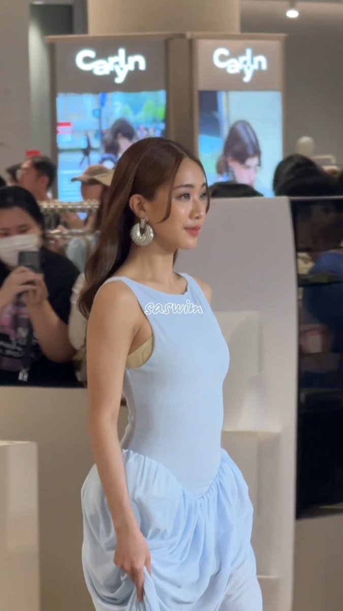 A young woman with long brown hair wearing a light blue sleeveless dress and silver hoop earrings poses at what appears to be a 'Carlyn' branded store event, with the watermark 'saswiia' on the image.