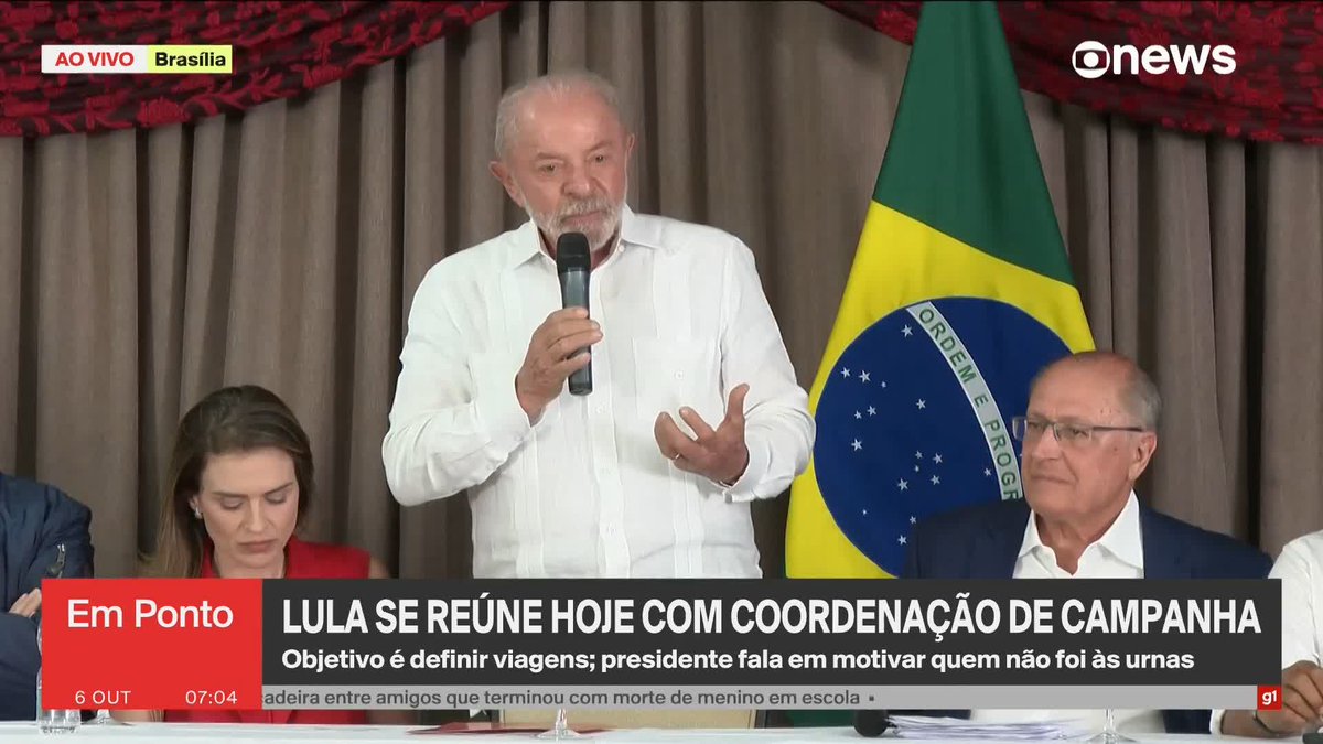President Luiz Inácio Lula da Silva, flanked by a Brazilian flag and seated allies, speaks into a microphone during a live GloboNews broadcast from Brasília on October 6, 7:04 AM, addressing his campaign coordinators about defining travel plans and motivating the 30 million Brazilians who abstained from voting.