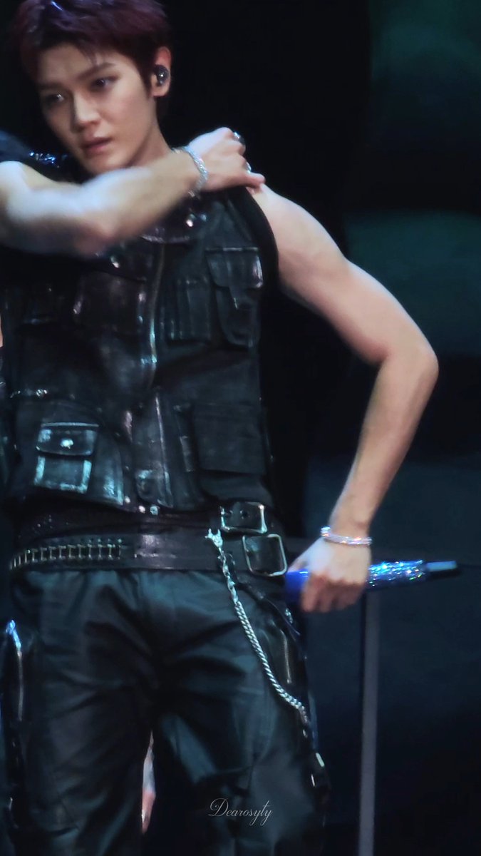 Stage performance photo of a K-pop performer with reddish-purple hair wearing a black leather utility vest with chains and buckles, holding a blue microphone.
