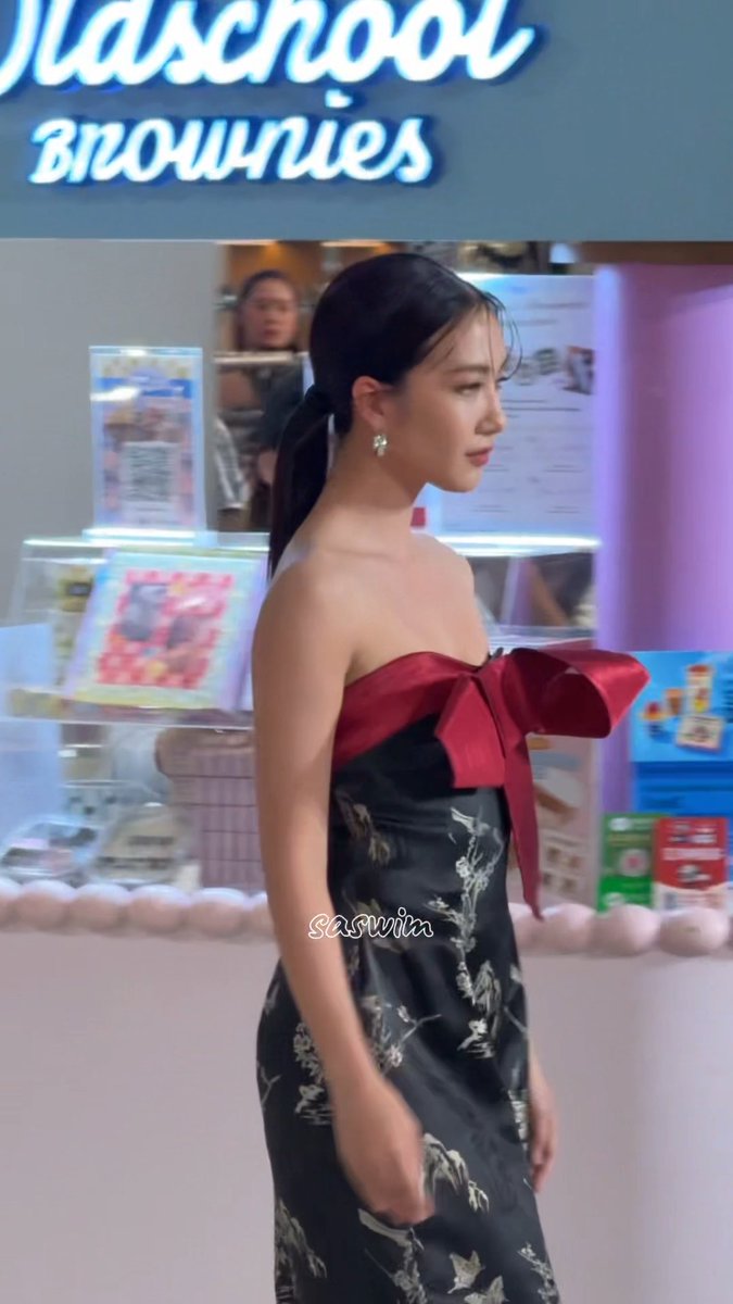 A woman in a strapless black floral dress with a large red bow standing in front of a 'Oldschool Brownies' storefront, with the watermark 'saswia' visible.