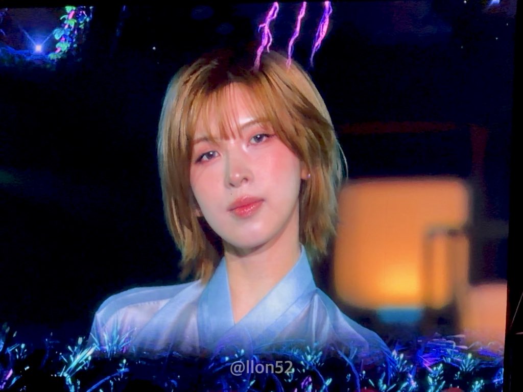 An artistic photo shows a person with short blonde hair and a light blue sheer top, surrounded by blue and pink floral elements and lights, with the watermark '@llon52'.