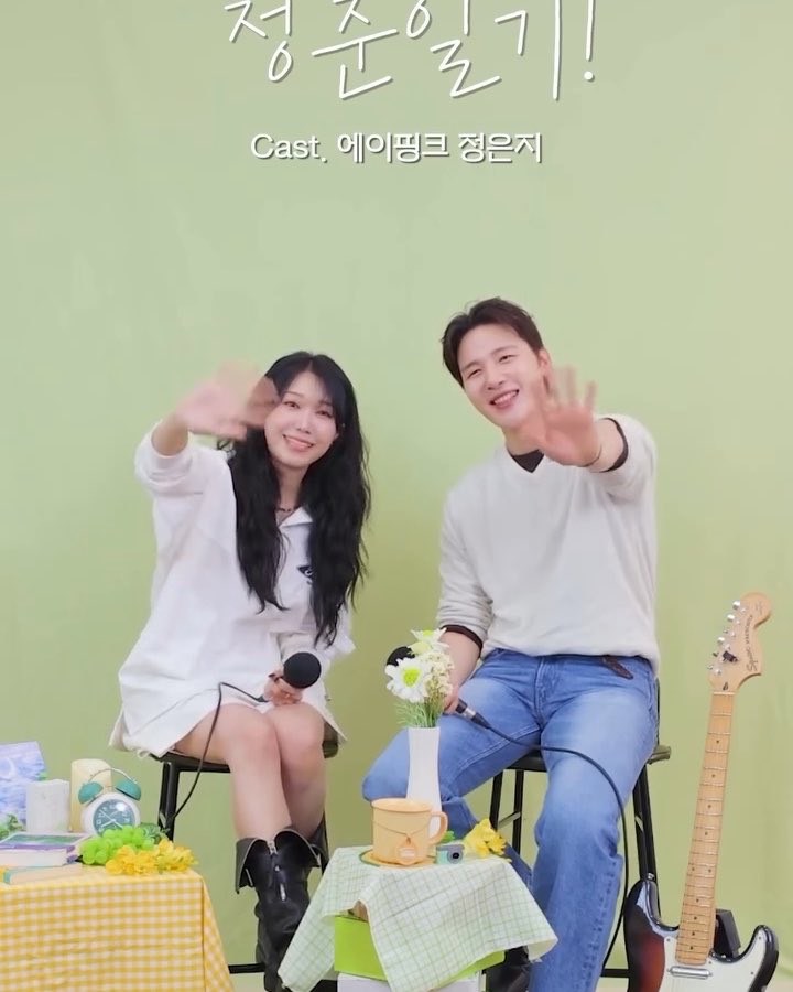 Two smiling young people, a man and a woman, sitting on stools and waving at the camera in what appears to be a Korean variety show setting, with Korean text at the top and a guitar visible on the right.