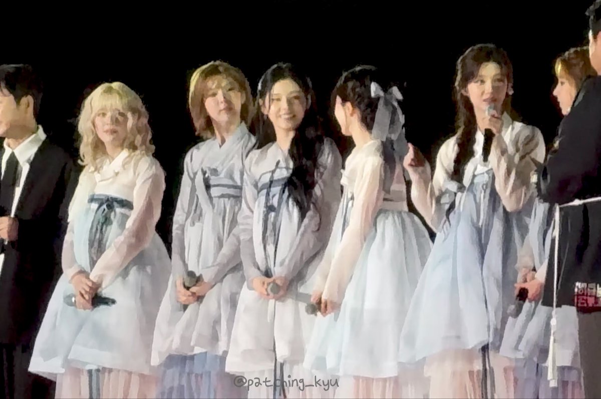 A group of performers on a dimly lit stage wearing light blue and white traditional-style dresses, holding microphones, with a watermark reading '@patching_kyu'.