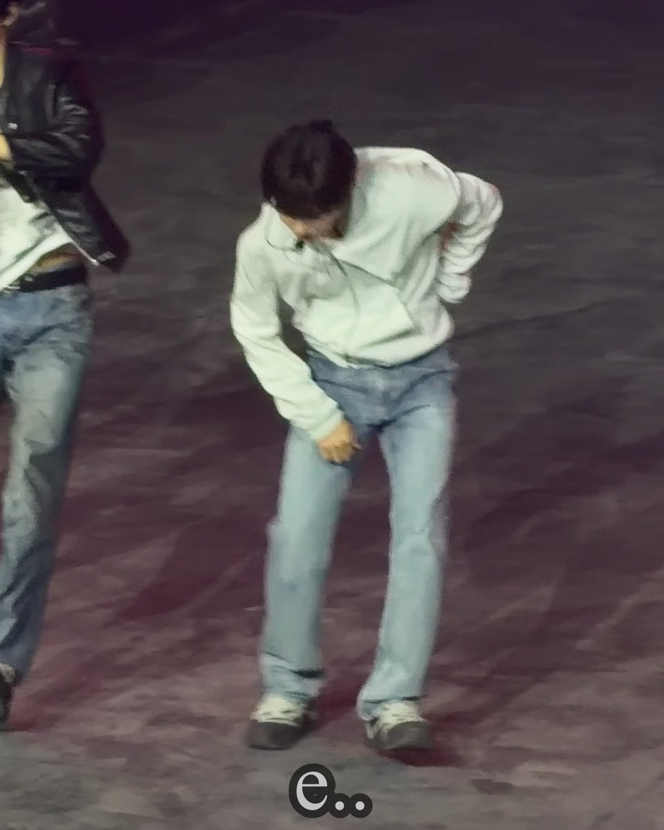 A person in a white hoodie and light blue jeans stands bent over in dim lighting on a paved surface, with another person partially visible on the left and a watermark at the bottom.