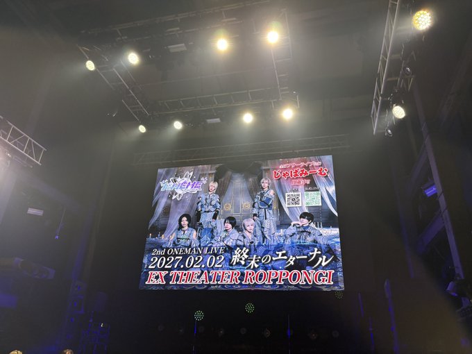 A concert venue stage screen displaying an advertisement for "2nd ONEMANLIVE 2027.02.02 終末のエチュード EX THEATER ROPPONGI" with images of performers and a QR code, lit by overhead stage lamps.