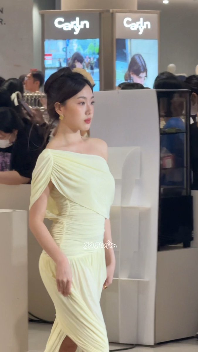 A woman in an elegant off-shoulder pale yellow dress poses at an event, with screens displaying 'Caryn' logos in the background and 'sasviiix' watermark visible.