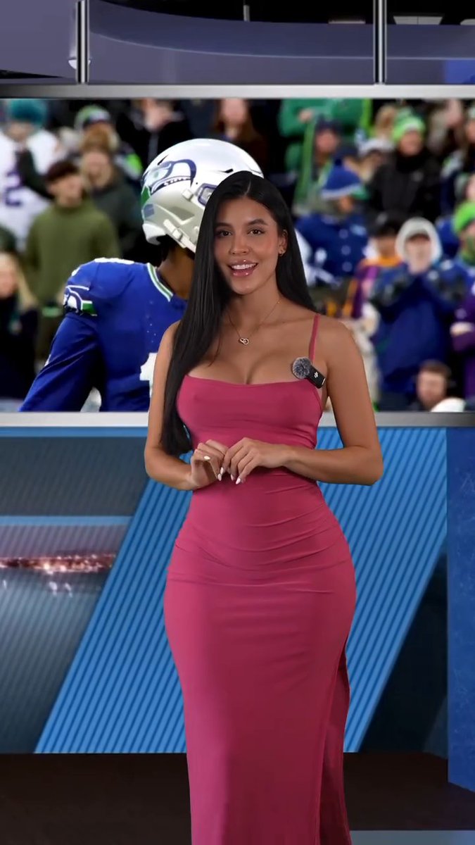 A woman with long black hair wearing a form-fitting pink dress stands smiling on a broadcast set with a Seattle Seahawks player and fans visible on a screen behind her.