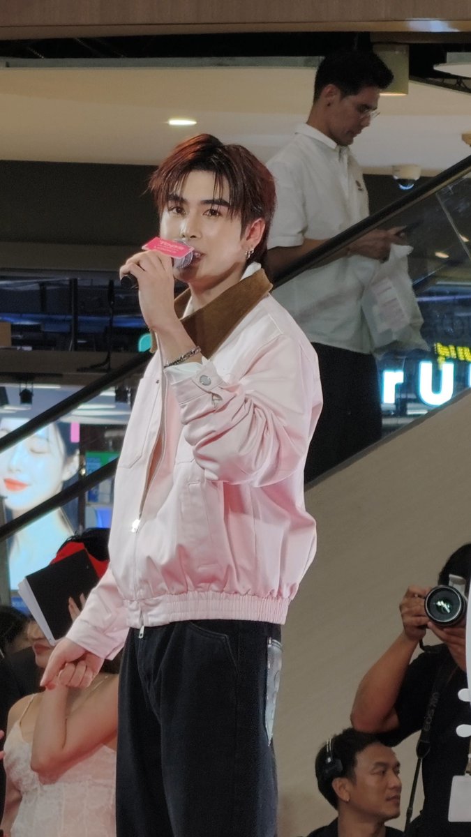 A young man with reddish-brown hair wearing a pink and white jacket speaks into a microphone at what appears to be a public event, with photographers and onlookers in the background.