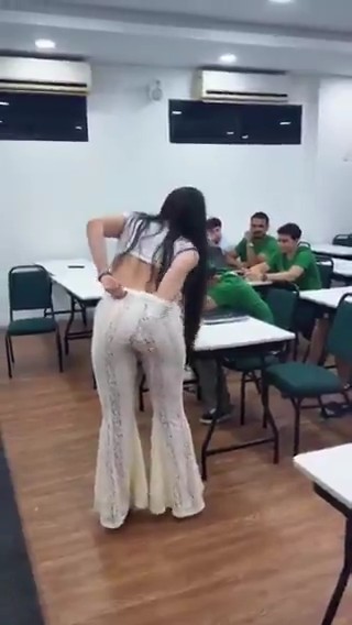 A blurry video still showing a woman with long dark hair wearing white pants and a black and white top, standing in a room with men seated at tables in the background.