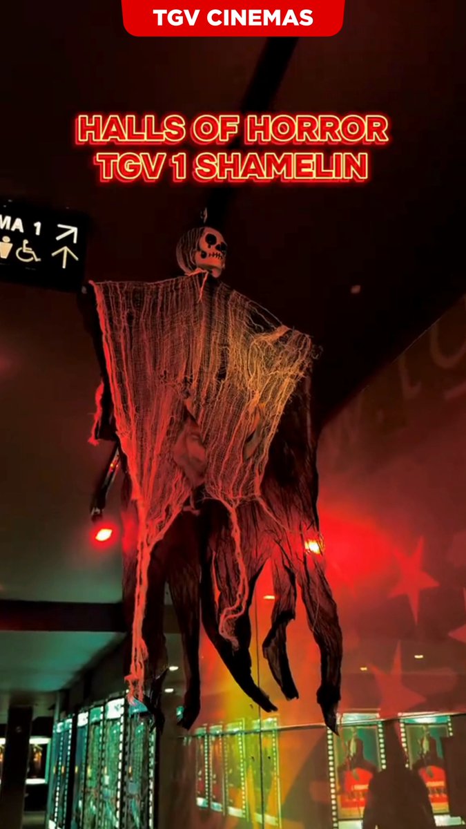 A Halloween decoration at TGV Cinemas showing 'HALLS OF HORROR TGV 1 SHAMELIN' with a hanging skeleton figure draped in tattered fabric illuminated by red lighting.