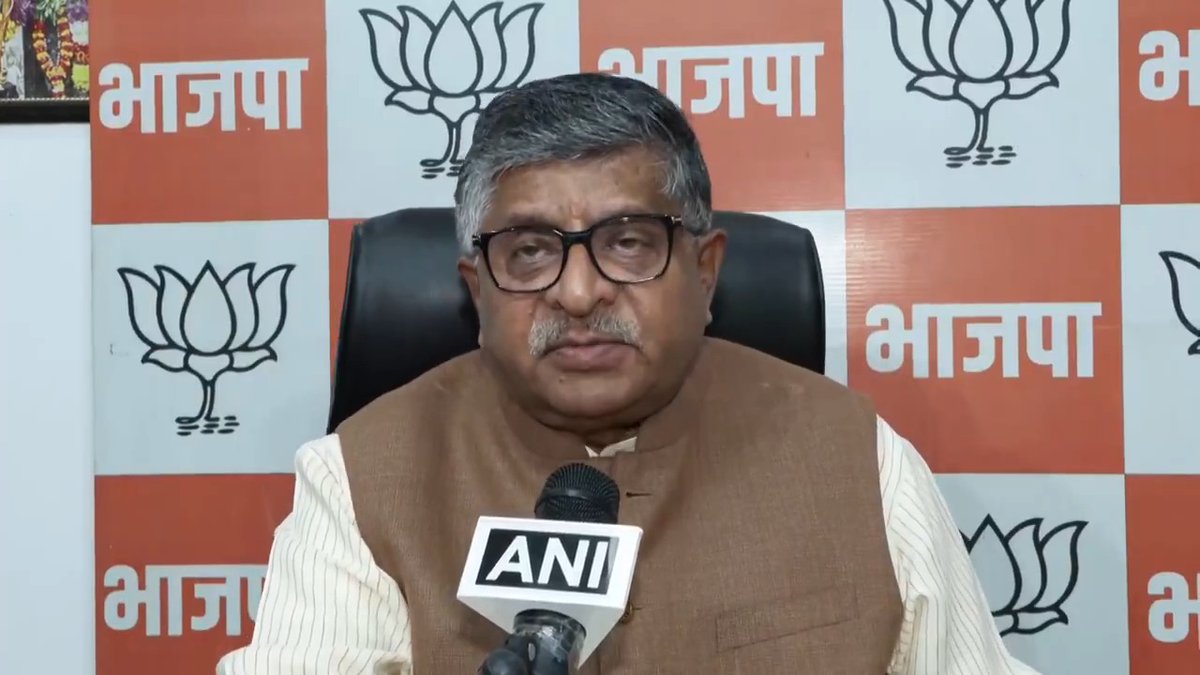 A man with glasses, wearing a brown vest over a striped shirt, speaks into an "ANI" microphone during a press conference. The backdrop features the lotus symbol and the word "भाजपा" (BJP) repeated, indicating he is a Bharatiya Janata Party representative commenting on the opposition's actions.
