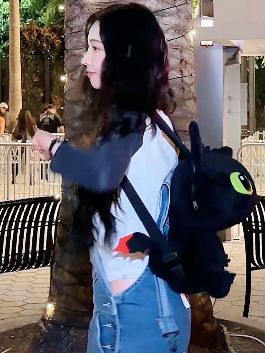 A woman with long dark hair is seen from the side wearing a light blue top and jeans, carrying a large black Stitch backpack at what appears to be a theme park.