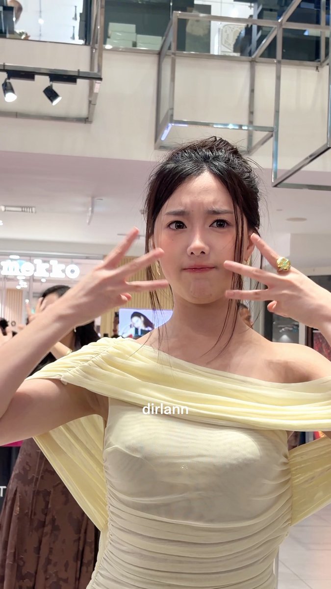 A young woman in a pale yellow off-shoulder draped dress makes a playful, pouting expression while holding up her hands in claw-like gestures inside a mall; a 'mekko' storefront sign is visible in the background, and the watermark 'dirlann' appears on the image.