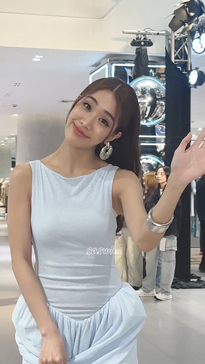 A young woman with long dark hair wearing a light blue sleeveless top and white skirt, posing at what appears to be a public event with stage lighting rigs visible behind her. The watermark 'saswiix' is visible on her clothing.