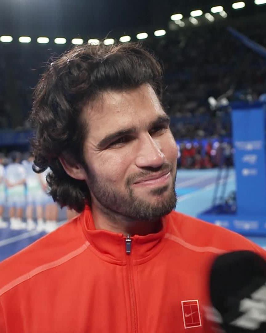 A close-up image shows tennis player Carlos Alcaraz being interviewed, wearing a red Nike zip-up jacket featuring a tennis racket logo, with a stadium crowd blurred in the background. The setting reflects his post-match press moment following his back-to-back championship win in Tokyo, as mentioned in the caption.