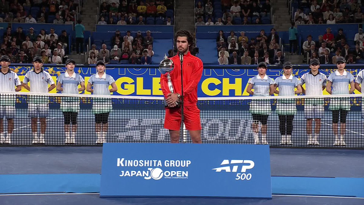 Carlos Alcaraz stands on court holding the championship trophy at the Kinoshita Group Japan Open Tennis Championships (ATP 500), surrounded by ball kids lined up behind the net during the trophy ceremony following his first title win since February.