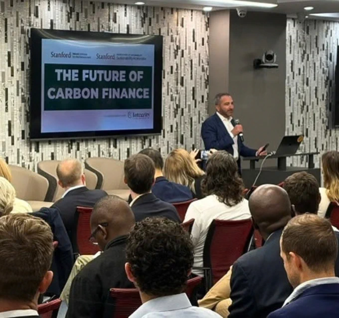 A speaker holds a mic in front of an audience next to a slide that says, The future of carbon finance