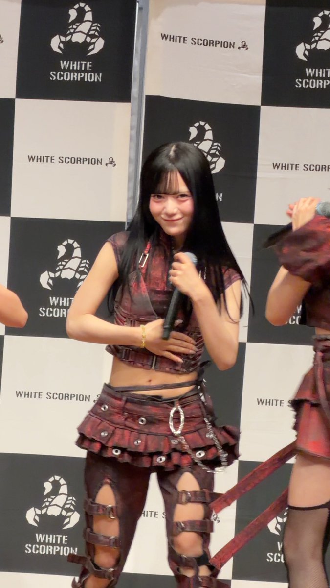 A performer with long dark hair smiles at the camera while holding a microphone, wearing a distressed red plaid top, a matching skirt, and cutout leggings against a 'WHITE SCORPION' logo wall.