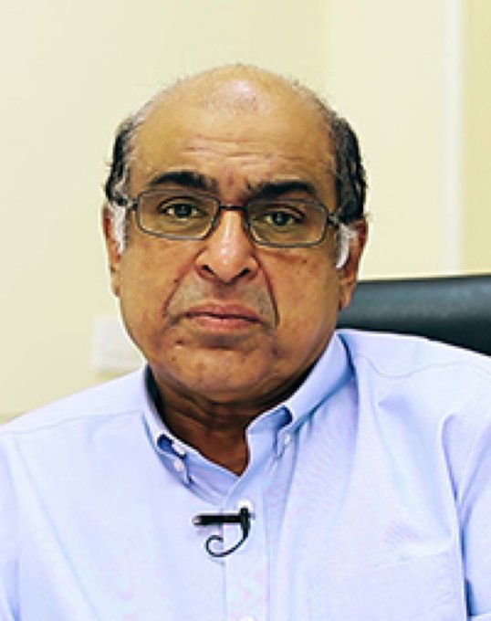 A headshot of an older bald man wearing glasses and a light blue button-up shirt with a small microphone clipped to it.