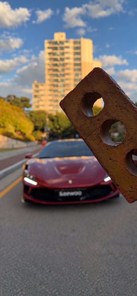 A red Ferrari with 'Saewood' on the license plate drives down a road, with a rusty metal bracket featuring circular cutouts held up in the foreground.