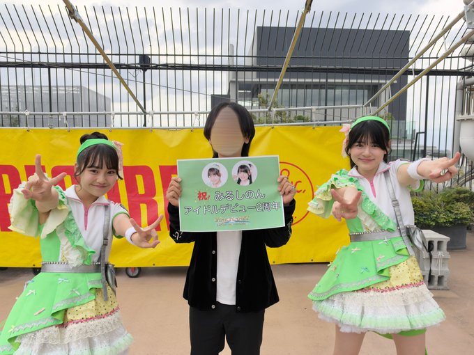 Two young women in green and white frilly outfits pose next to a person holding a sign that reads '祝 みるしぃのん アイドルデビュー2周年' (Celebrating Mirushion's 2nd Anniversary of Idol Debut).
