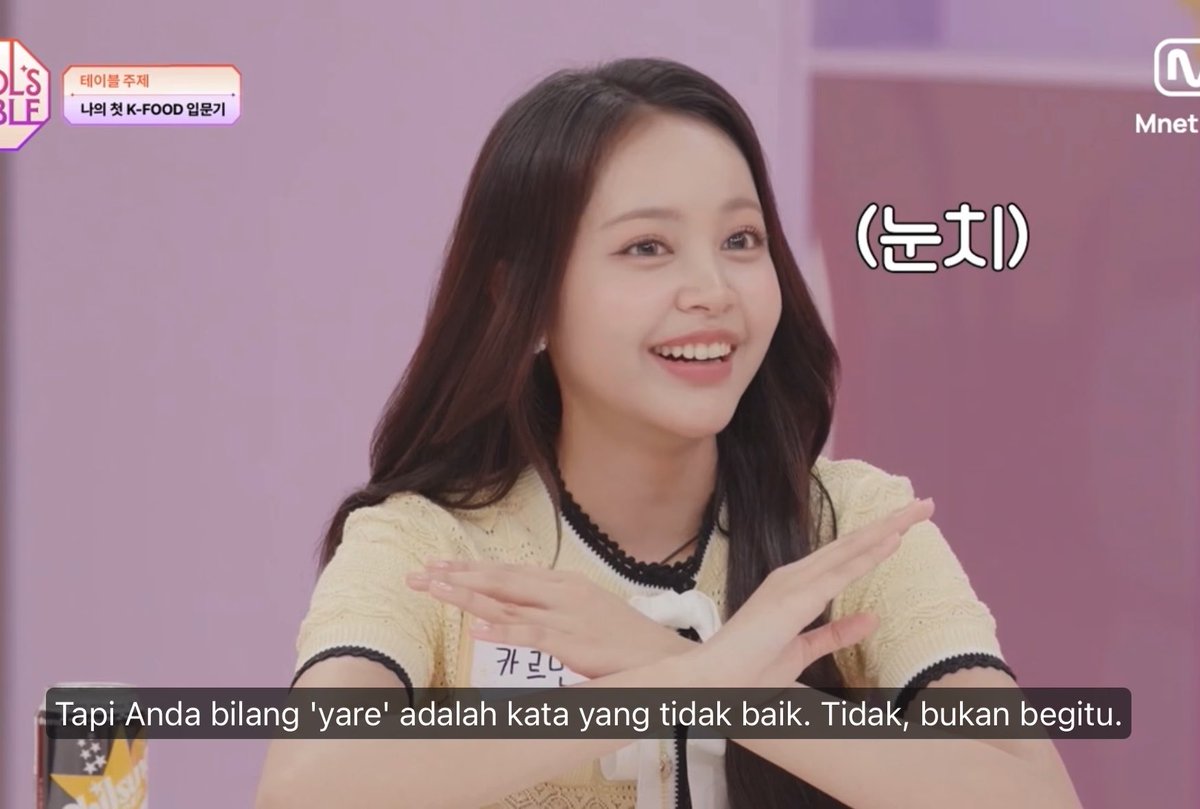 Screenshot from an Mnet K-pop show with Indonesian subtitles, showing a smiling female idol crossing her arms, with on-screen text "(눈치)" and "Tapi Anda bilang 'yare' adalah kata yang tidak baik. Tidak, bukan begitu."