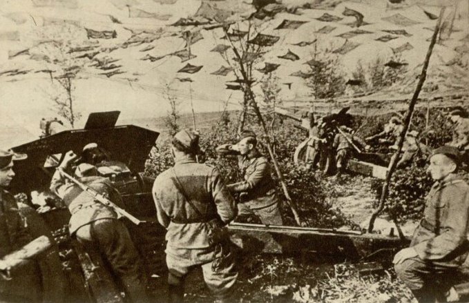 A vintage black and white photograph showing soldiers with rifles positioned in a mountainous, hilly terrain, with what appears to be artillery in the foreground.