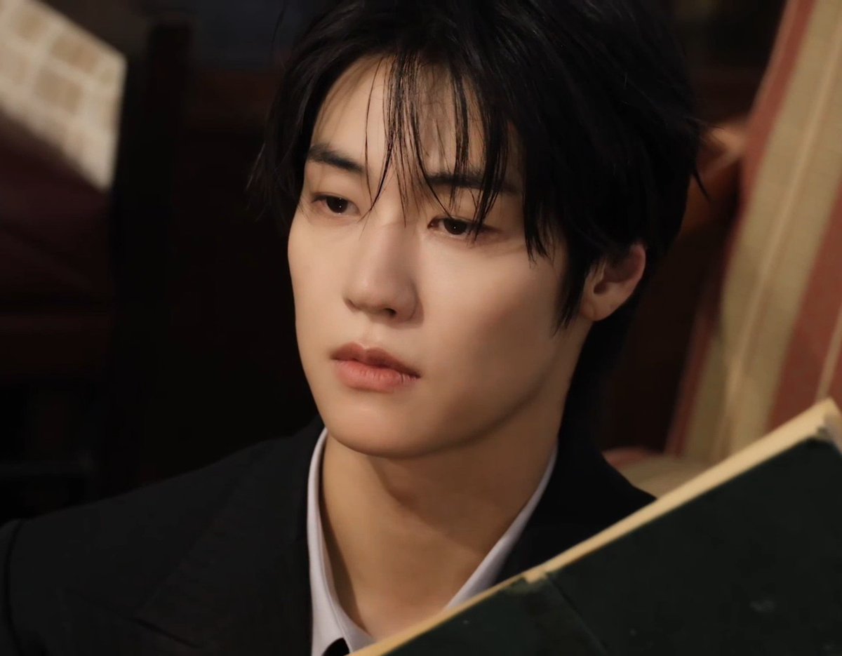 A close-up portrait of a young man with dark, wet-looking hair wearing a black blazer over a white shirt, looking off to the side while holding a dark object.