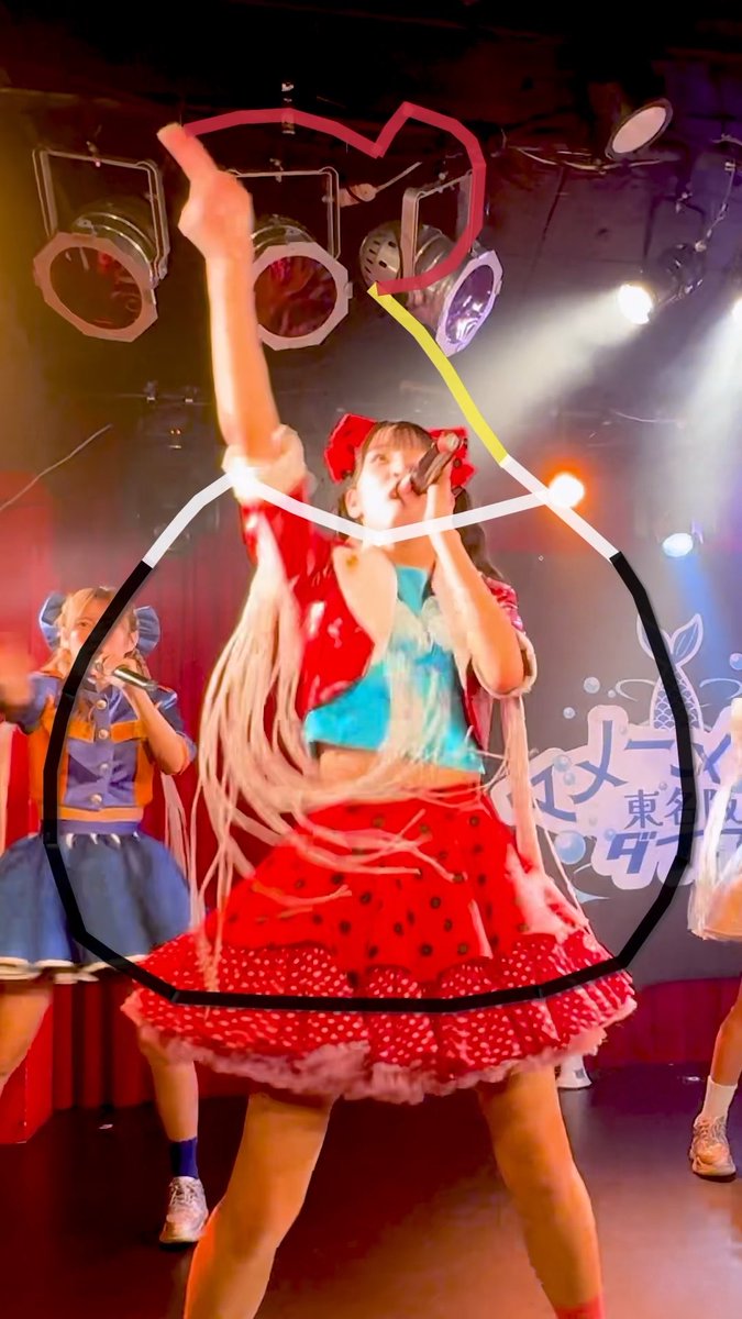 A performer in a red polka-dot dress with blue top performing on stage, overlaid with a cartoon bomb outline drawn in pink and yellow.